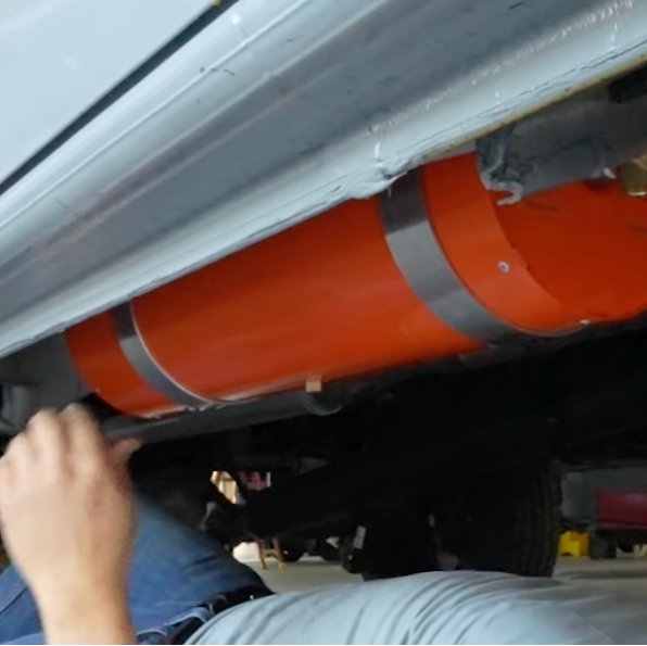 An INCLINE WATER HEATER  mounted beneath a Sprinter, with a person's hand and lower body visible working underneath in a garage or workshop setting.