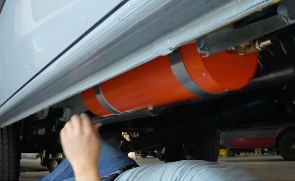 An INCLINE WATER HEATER  mounted beneath a Sprinter, with a person's hand and lower body visible working underneath in a garage or workshop setting.